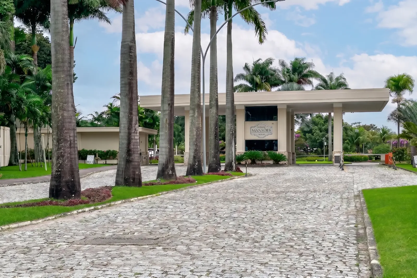 Portaria principal do Condomínio Mansões Barra da Tijuca — acesso com palmeiras imperiais e guarita blindada